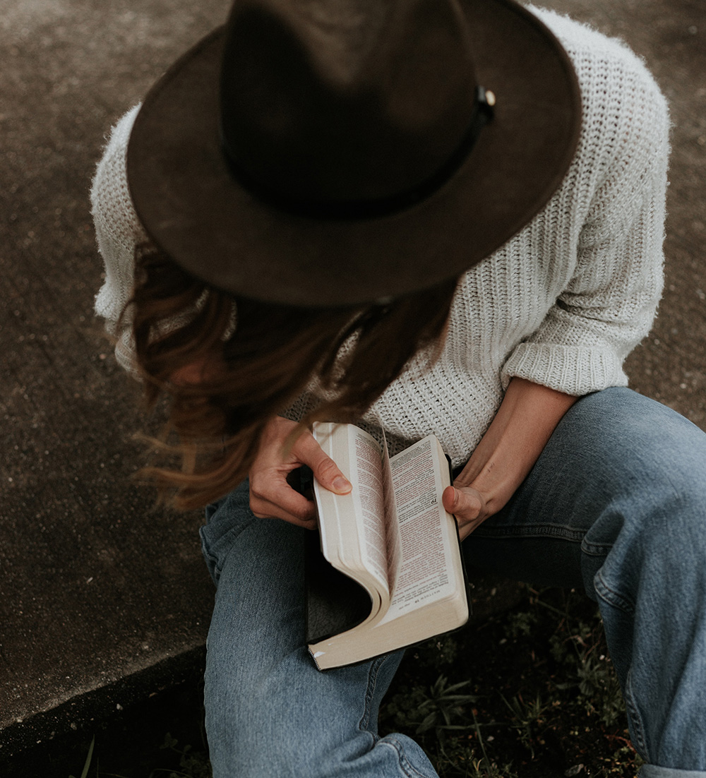 woman wearing a stylish brown hat sitting on a curb opening her Bible